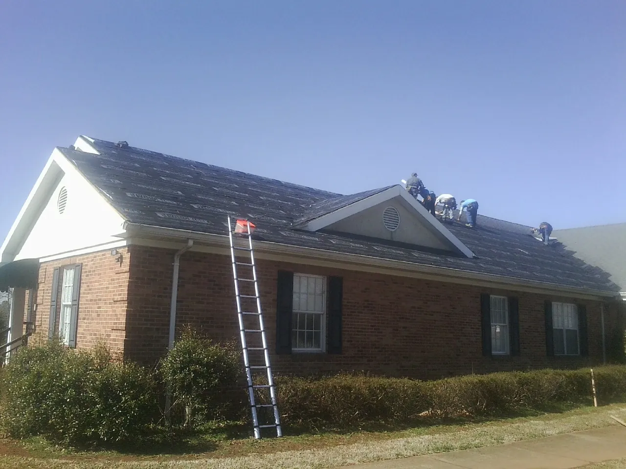 Crew tearing shingles off a brick ranch roof with a ladder set — Emergency Roof Repair work by our crew in Stirum