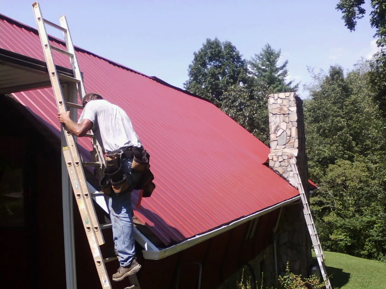 Roofer on a ladder beside a red metal roof and stone chimney — Soffit & Fascia Repair by Roofing Brothers in Stirum